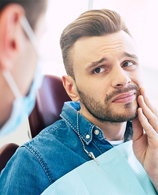 Man in dental chair looking at dentist holding hand to jaw in pain